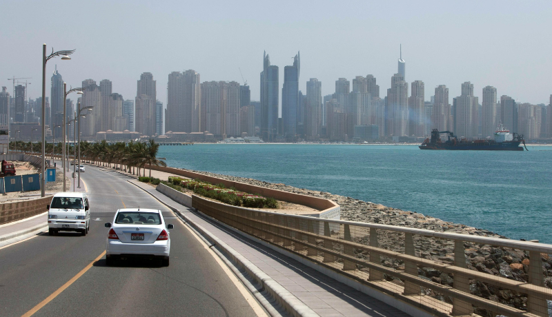 A coastal drive beside calm blue water with a Dubai city full of high-rise towers in the background.