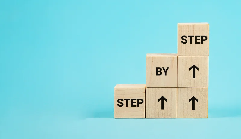 Wooden blocks arranged like stairs with the words step by step on a blue background.
